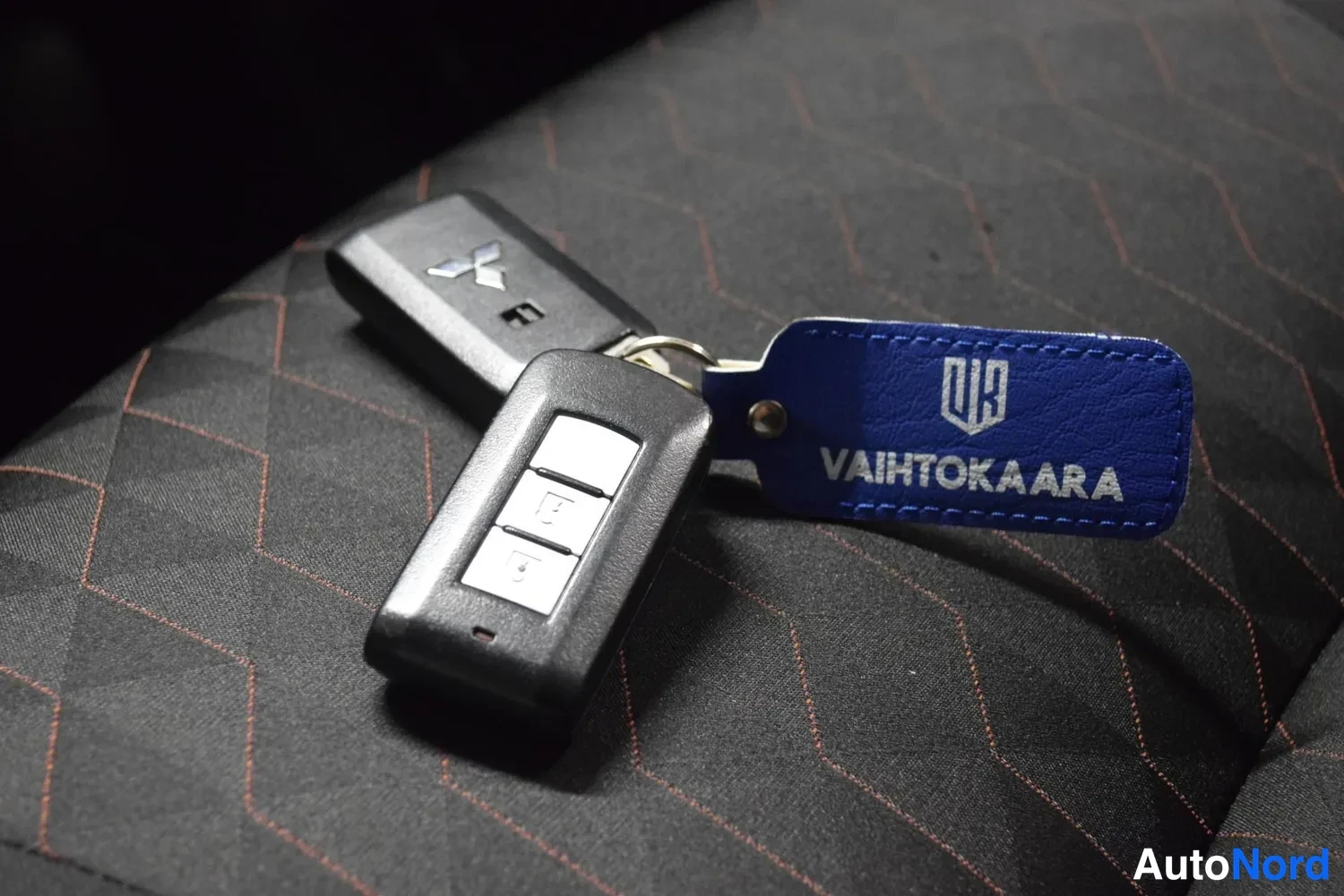 Yksityiskohta: Kaksi Mitsubishi avainlokeroa avaimenperässä sinisellä VAIHTOKAARA-avainnauhalla kuviollisen istuinkankaan päällä.