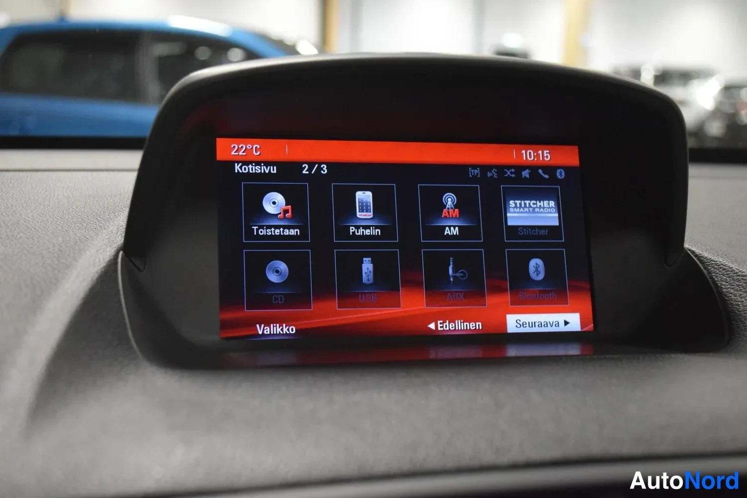 Muu: Infotainment-näytön Kotisivu-valikko (2/3) kuvakkeineen: Toistetaan, Puhelin, AM, Stitcher, CD, USB, AUX ja Bluetooth; kellonaika 10.15, lämpötila 22 °C.