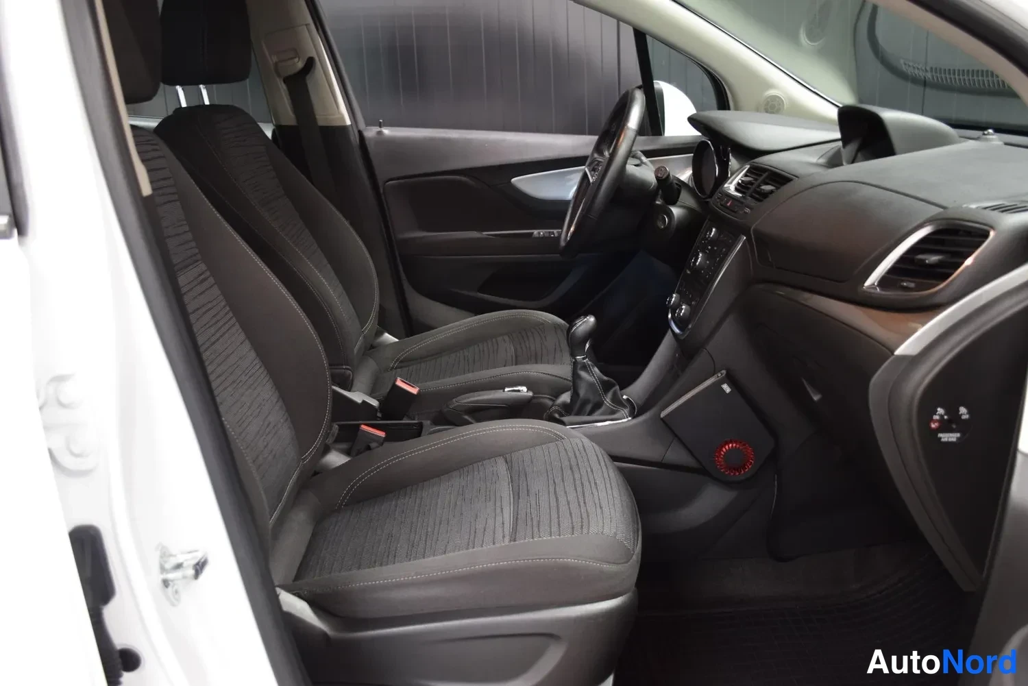Muu: auto sisältä kuvattuna kuljettajan oven puolelta: tummat kankaat etuistuimet, manuaalivaihteen vipu ja kojelauta.