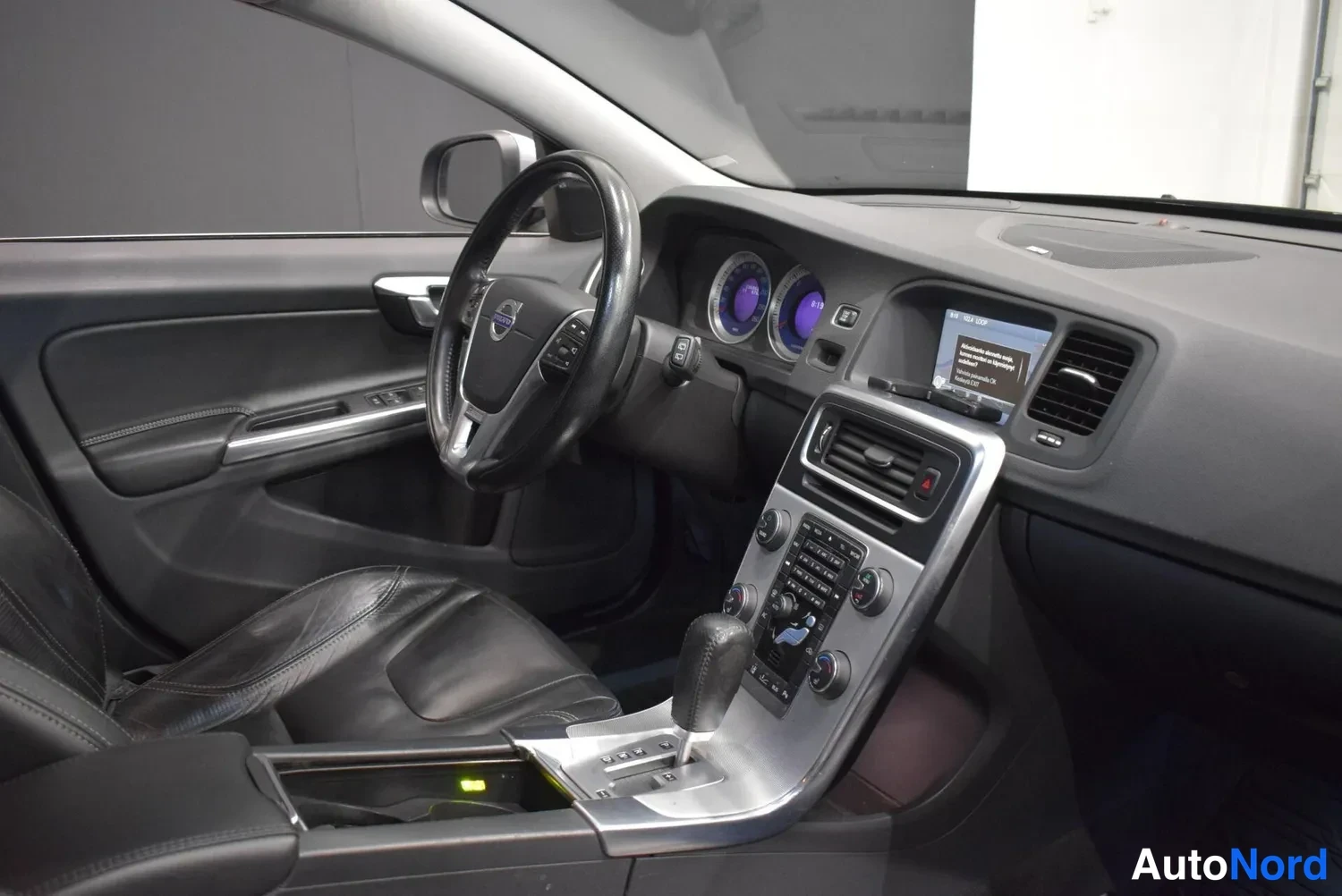 Muu: Volvo V60:n ohjaamo kuljettajan puolelta: nahkainen ohjauspyörä, siniset mittarit, keskikonsoli ja automaattivaihteen valitsin, tummat verhoilut.