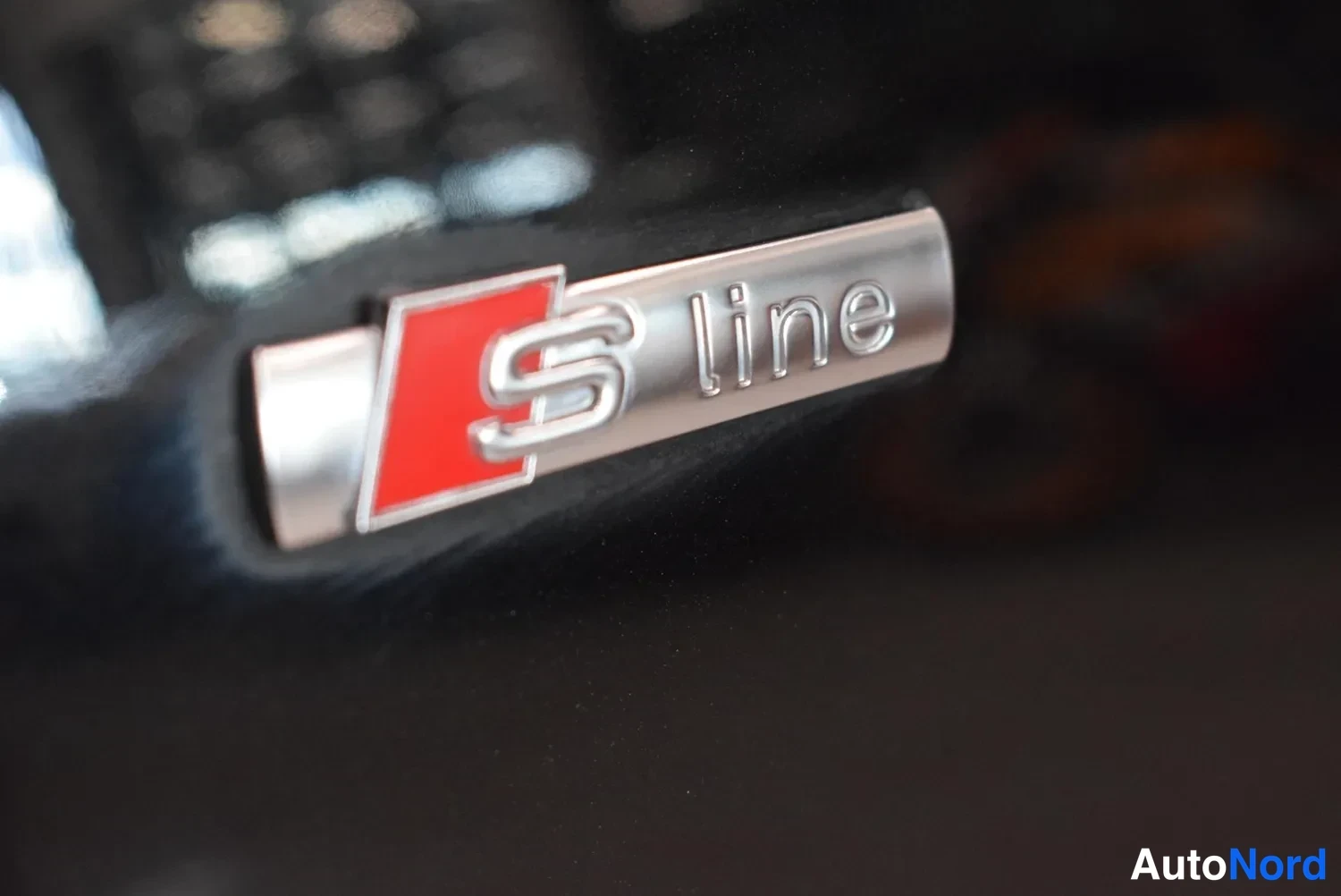 Yksityiskohta: Lähikuva mustan auton S line -merkistä punaisella S-logo metallisessa kilvessä.