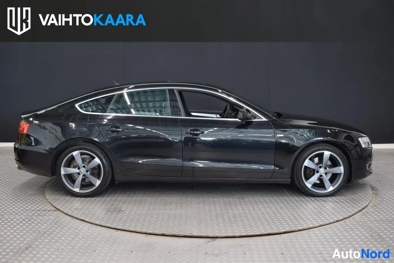Etuviisto, nokka oikealle: Mustan Audi A5 Sportbackin etukulma vasemmalta kuvattuna sisätiloissa alustalla.