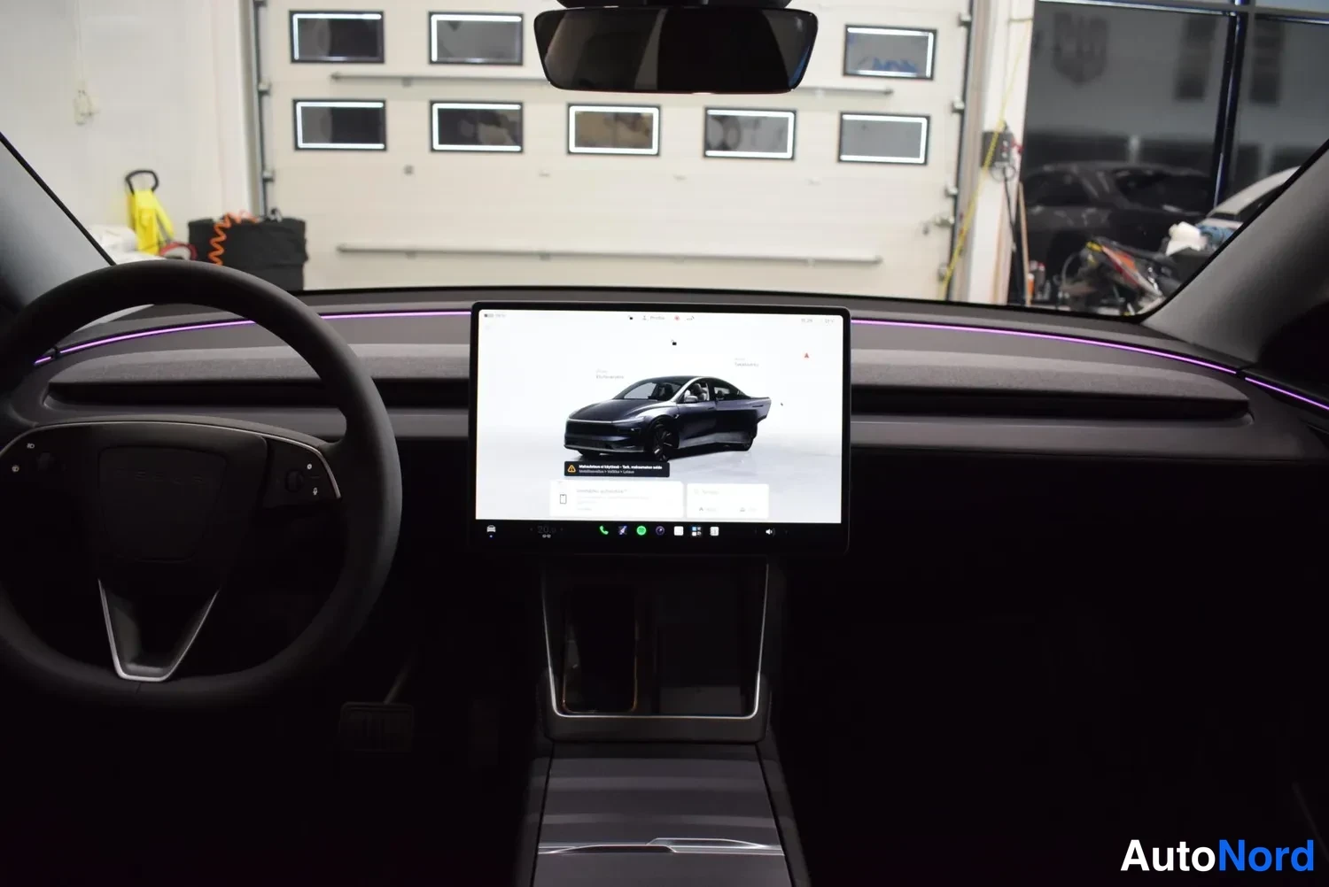 Yksityiskohta: Ohjaamo vasemmalta kuvattuna: puoliavoin steering wheel ja keskitason suuri kosketusnäyttö, jonka ruudulla auton kuva.