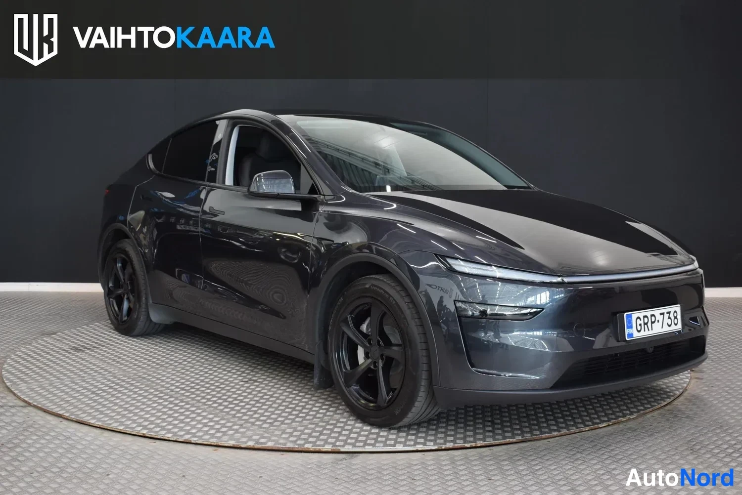 Etuviisto, nokka oikealle: Tesla Model Y -henkilöauto tummanharmaassa värissä etukulmasta oikealta kuvattuna studiotyylisessä tilassa.