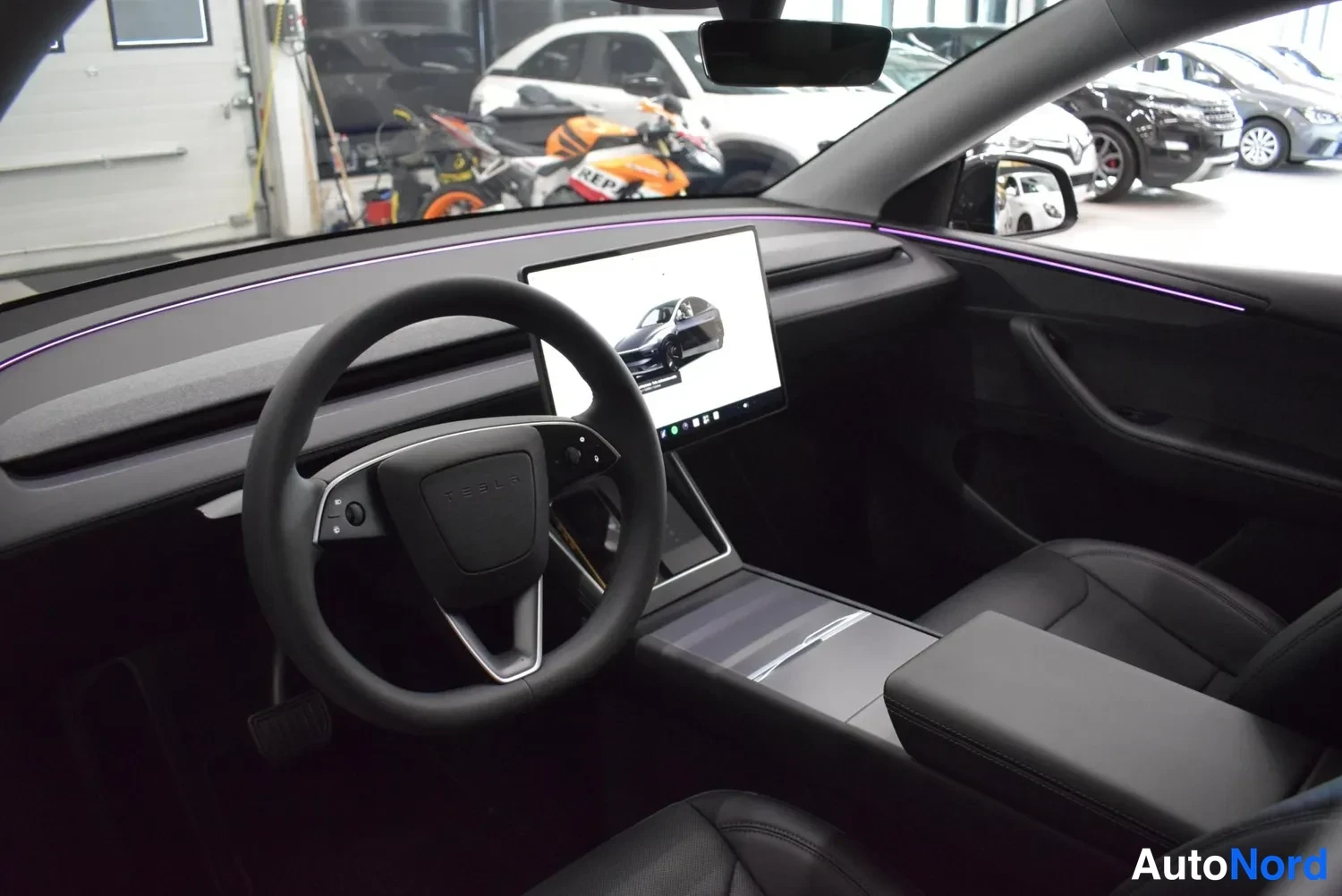 Muu: Autojen sisätilat ohjaamosta: Tesla-ohjauspyörä, keskikonsolin näyttö ja mustat penkit, taustalla autotallia ja moottoripyörä.