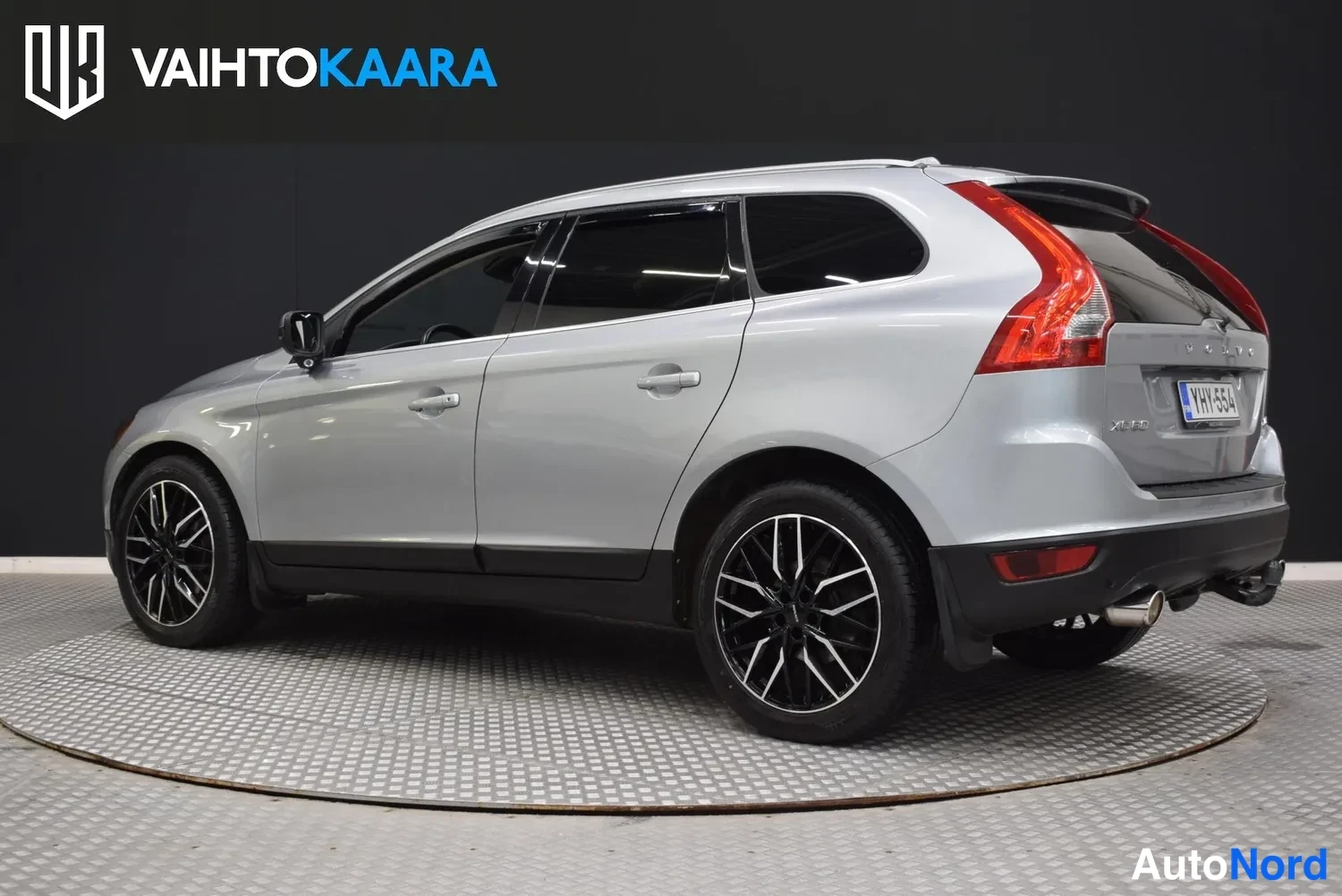 Takaviisto: Hopeanharmaan Volvo XC60:n takaokolikuskuva oikealta, rekisteritunnus, punaiset takavalot ja tummat vanteet.