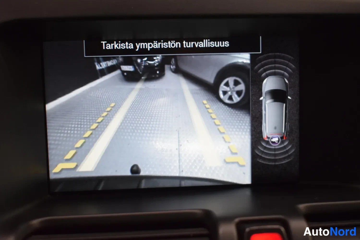 Yksityiskohta: Infotainment-näytöllä peruutuskameran kuva ja varoitus Tarkista ympäristön turvallisuus.