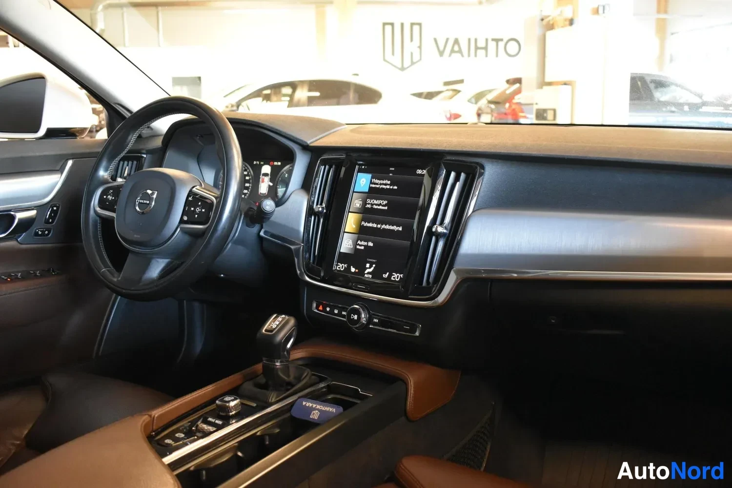 Sivulta: Volvo-ohjauspyörä ja tumma kojelauta, keskellä pystysuora kosketusnäyttö valikkoineen, keskikonsolissa vaihteenvalitsin ja ruskeat nahkaverhoukset.