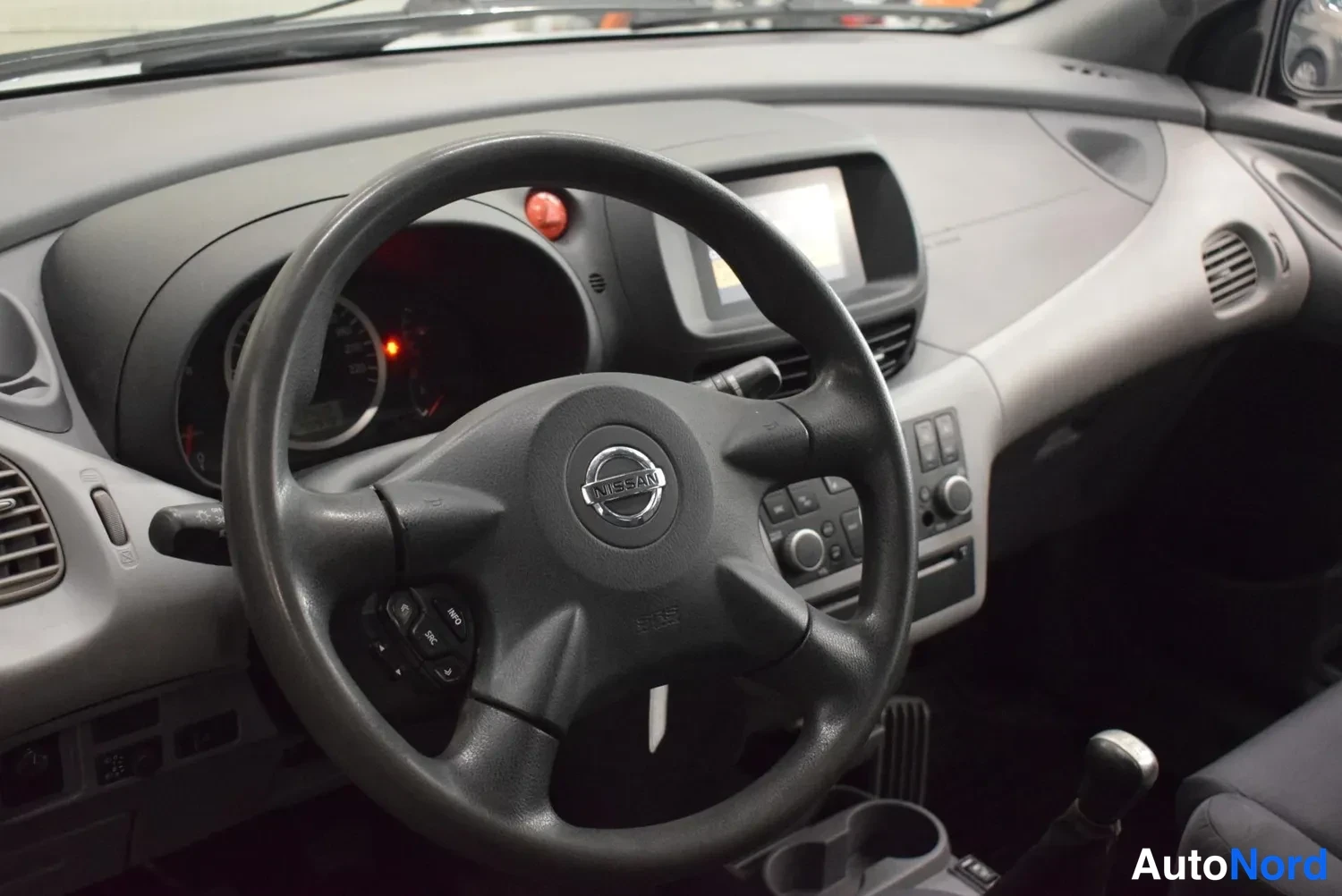 Muu: Ohjaamon näkymä Nissan-merkillä varustetun ohjauspyörän takaa: mittaristo, kojelaudan ruutu, radiopaneeli ja vaihtokeppi näkyvissä.