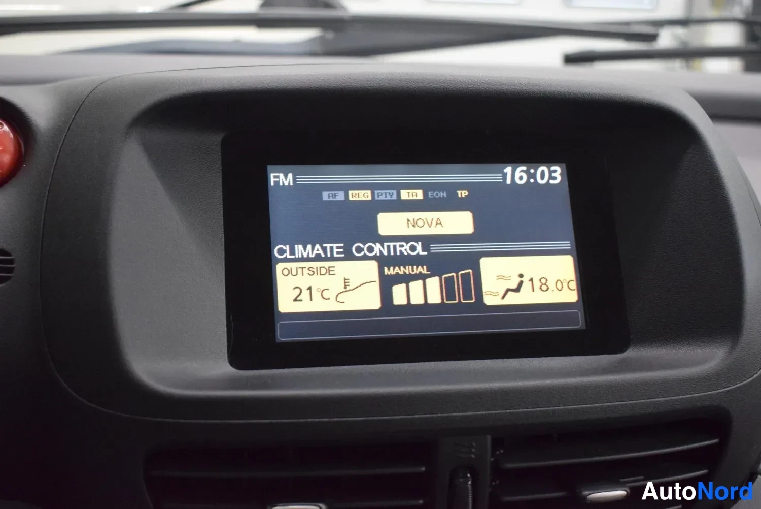 Yksityiskohta: Kojelaudan näyttö näyttää FM-radiota (NOVA), kellon aikaa 16:03 ja CLIMATE CONTROL -tiedot: ulkolämpötila 21°C ja manuaalinen ohjaus 18,0°C.