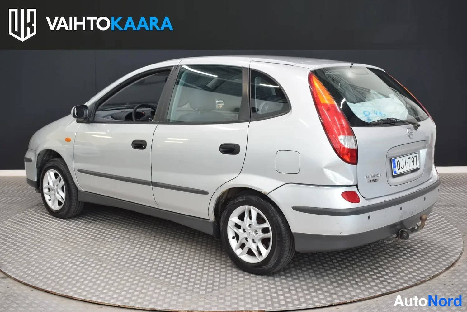 Takaviisto: Hopeanvärinen Nissan Almera Tino takaviistosta vasemmalta, rekisterikilpi.
