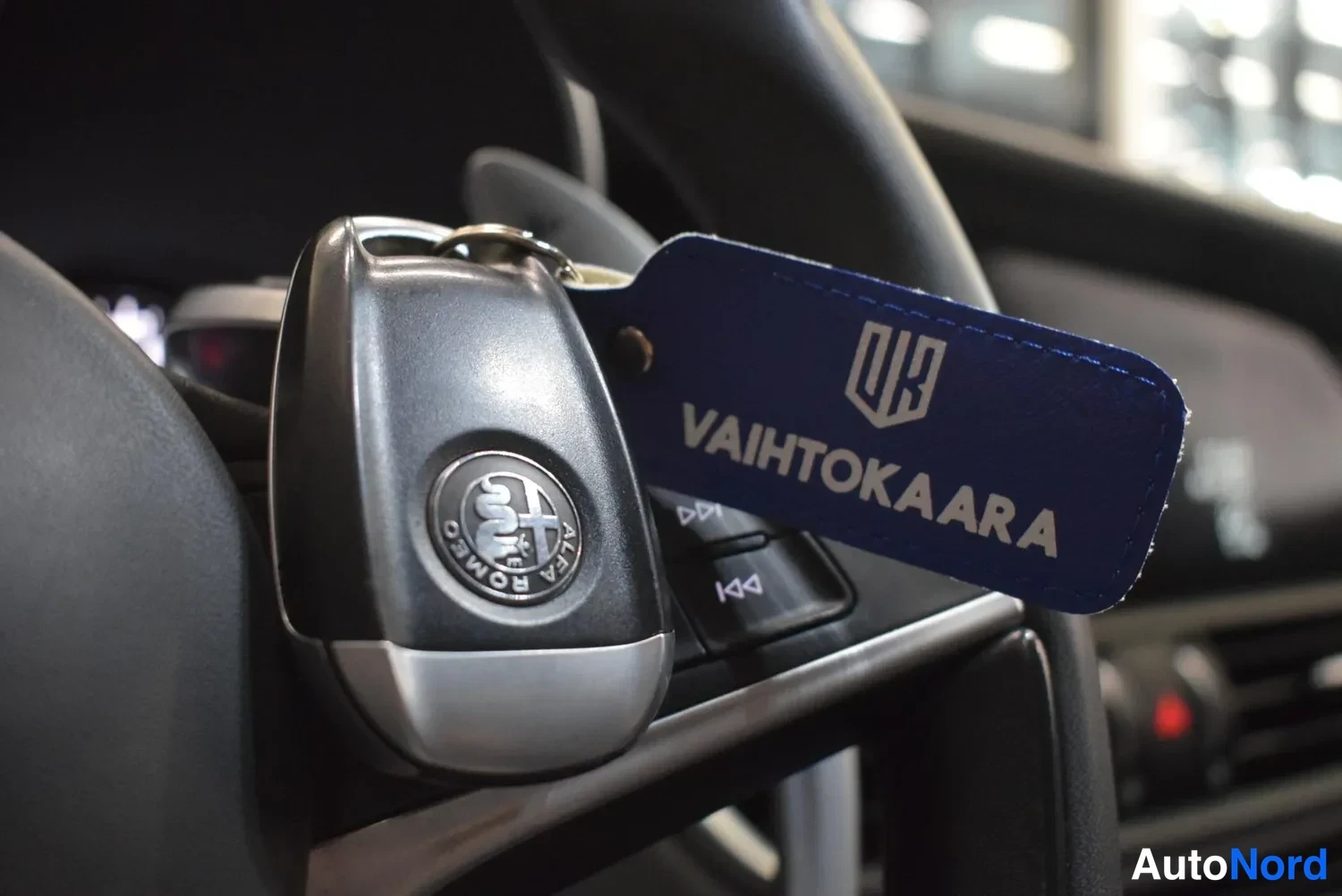 Yksityiskohta: Alfa Romeo -avainrimelli ja sininen Vaihtokaara-avainnauha ohjauspyörän近stä kojelautaa vasten.