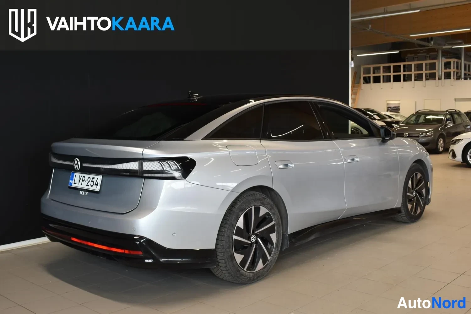 Takaviisto: Volkswagen ID.7 -sedanin takakulma vasemmalta, rekisterikilpi, hopea kori ja tumma lasikatto autosalissa.