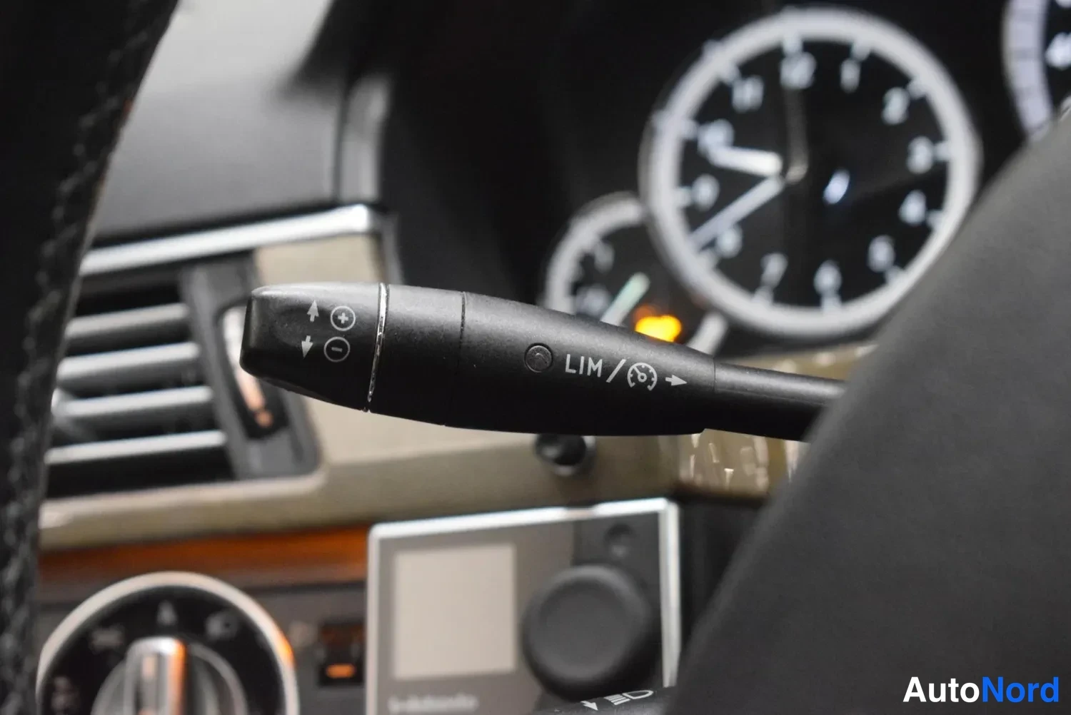 Yksityiskohta: Sisäkuva: Mercedes-Benzin ohjaustankytkin, jossa merkinnät LIM ja speedtronic-symboli sekä nuoli- ja plusmiinuspainikkeet, taustalla tumma mittaristo.