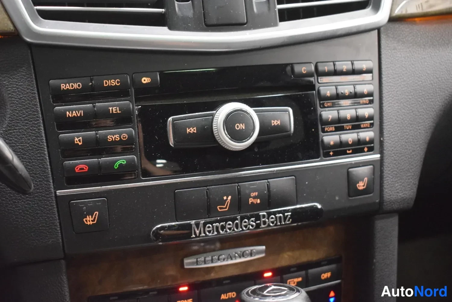 Yksityiskohta: Kojelaudan keskikonsolin lähikuva: radio-, NAVI-, TEL- ja puhelinpainikkeet, Mercedes-Benz-kyltti ja ELEGANCE-nimikilpi.