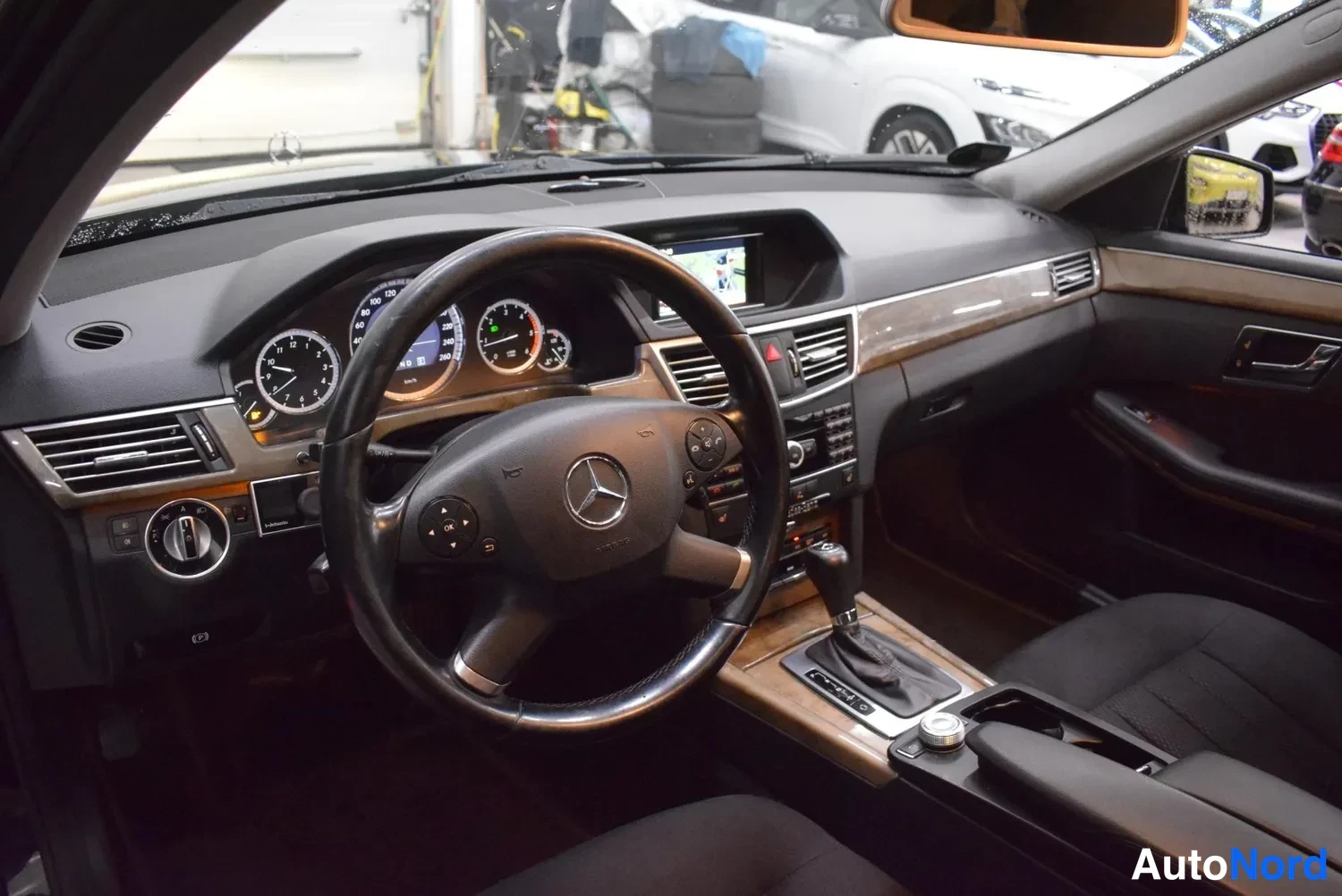 Muu: Auton ohjaamo etualalta: Mercedes-merkkiohjauspyörä, mittarit ja keskikonsoli, jonka näytöllä näkyy navigaatio.