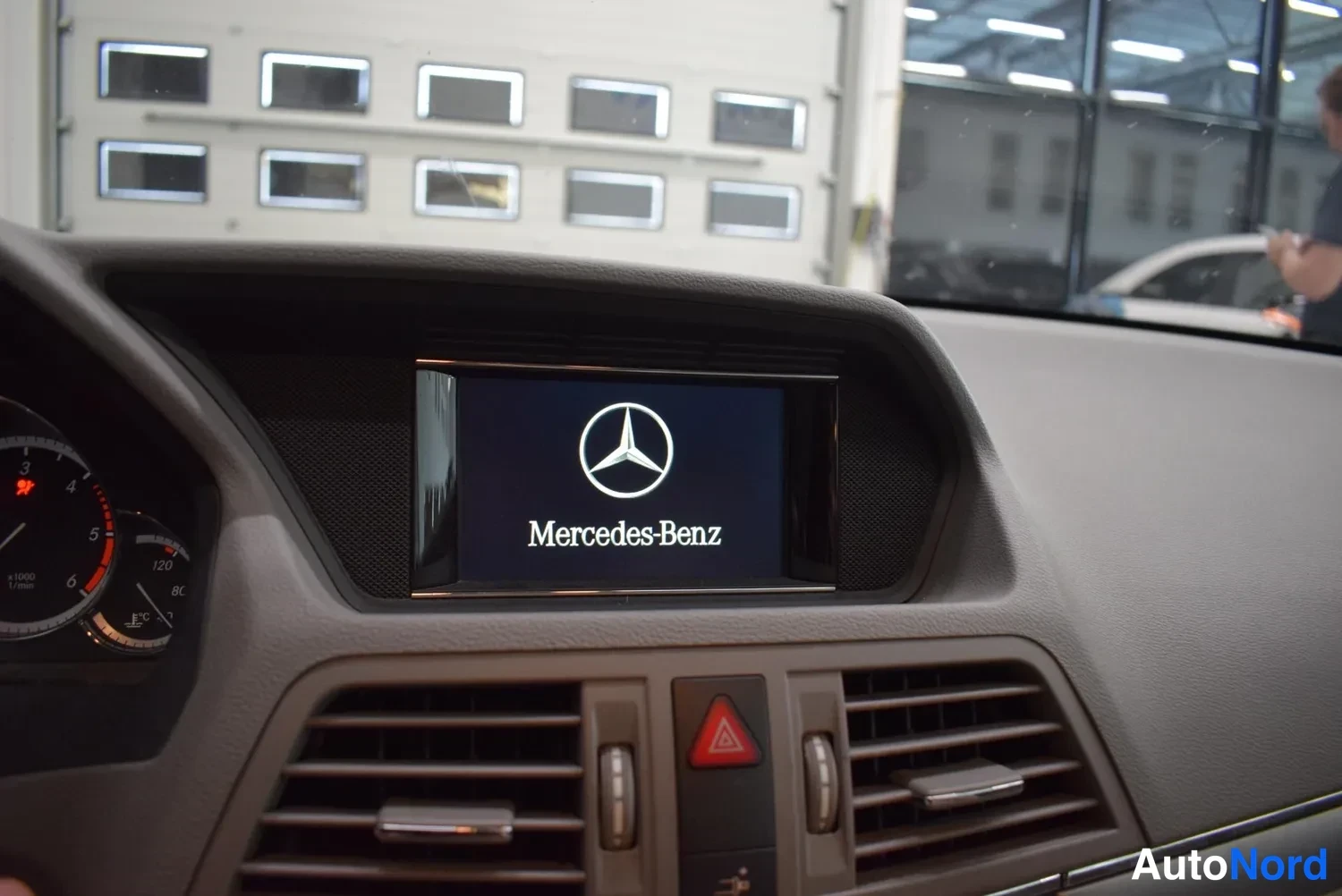 Yksityiskohta: Kojetaulun keskellä näyttö, jossa Mercedes-Benz-logo, vasemmalla osittain näkyvät rahjat ja mittarit.