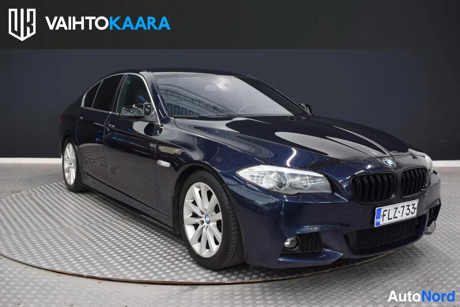 Etuviisto, nokka oikealle: Tummansininen BMW -sedan etukolmannesnäkymä oikealle kääntyneenä keulalla, rekisterikilpi.