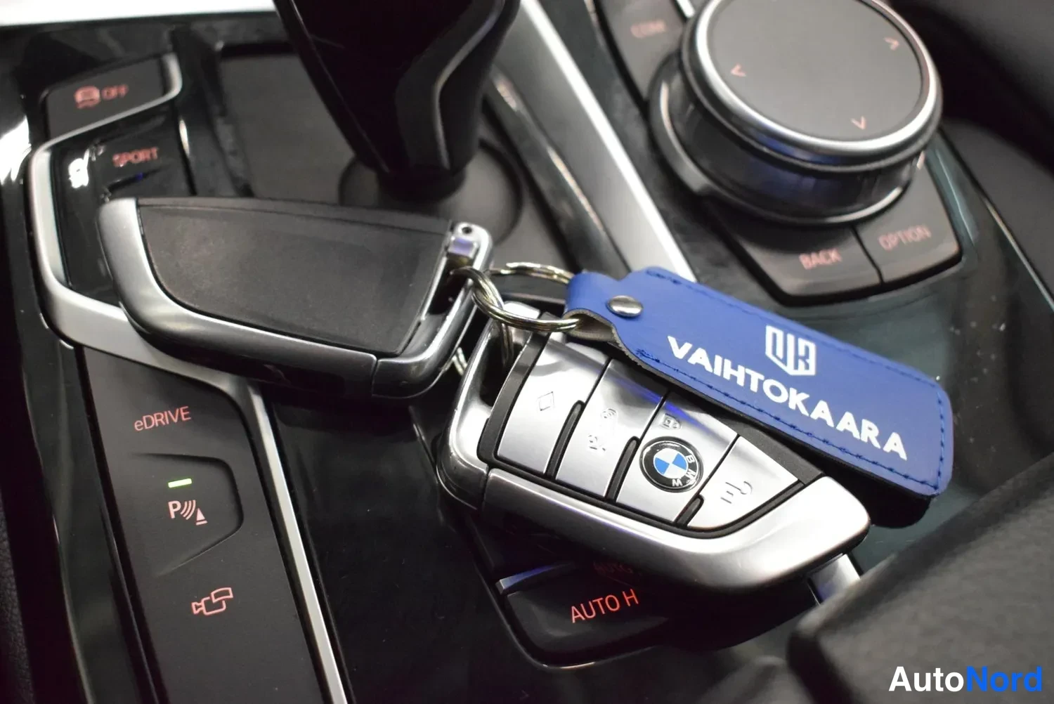 Muu: Keskikonsolin lähikuva: kaksi BMW-avainkätkintä avaimenperässä, jossa sininen VAIHTOKAARA-kuusi. Näkyvillä eDRIVE- ja AUTO H -painikkeet, iDrive-ohjain ja SPORT