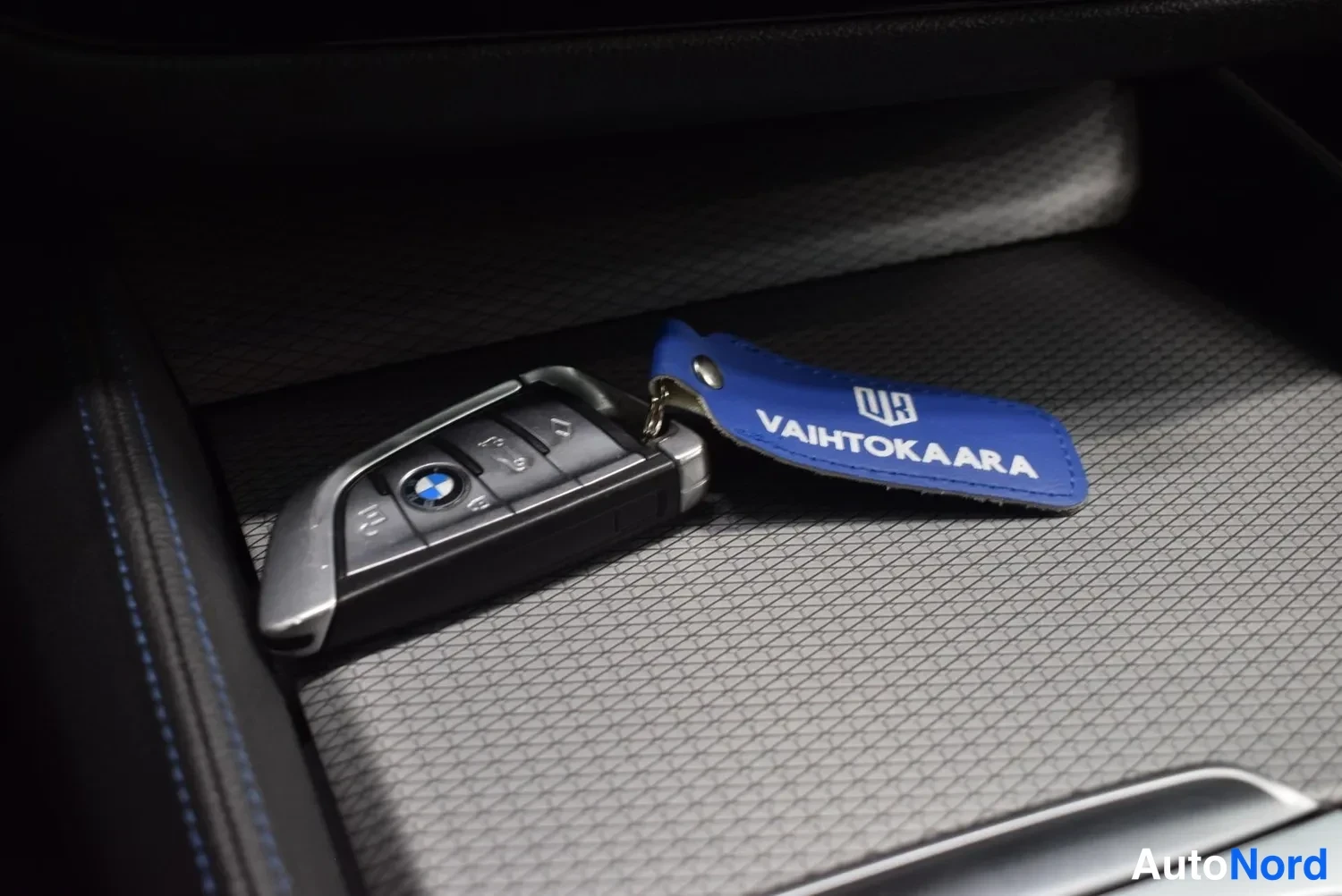 Yksityiskohta: BMW-avainavain ja sininen Vaihtokaara-avainnauha lokerossa, jossa on harmaa kuvioitu pohjamatto.