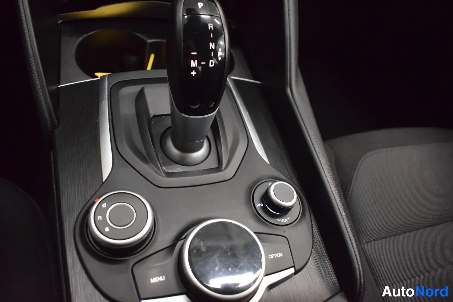 Yksityiskohta: Auton keskikonsoli automaattivaihteiston vaihdekeilalla, jonka merkit näyttävät P, R, N, D ja M-tekstit, sekä infotainment-järjestelmän MENU-, OPTION- ja volyym