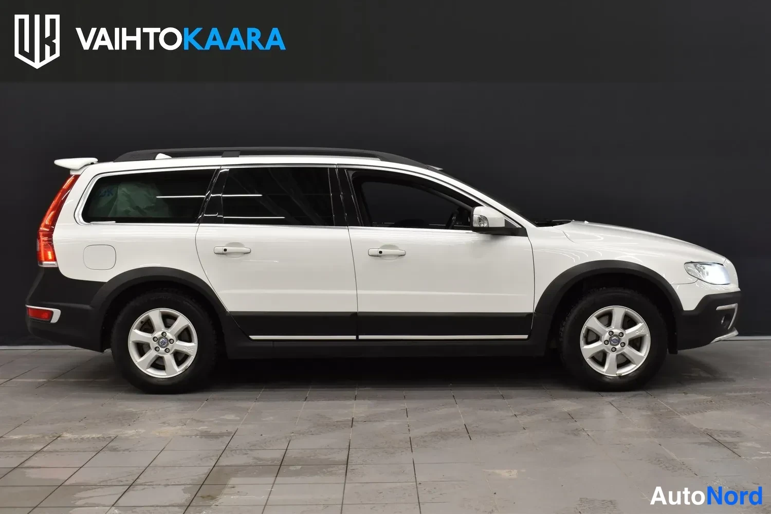 Sivulta: Volvo XC70 -farmariauto sivusta kuvattuna; valkoinen kori, mustat muoviosat ja kattokaiteet näkyvät.