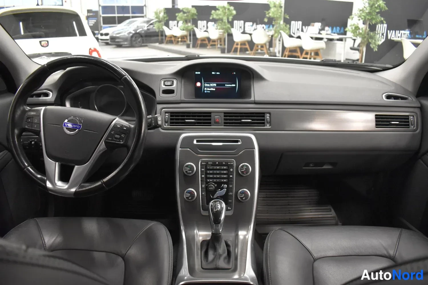 Muu: Volvo XC70:n ohjaamo sisältä: ohjauspyörä, kojelauta, keskitason infotainmentnäyttö ja automaattivaihteen valitsin, tummat nahkaistuimet.