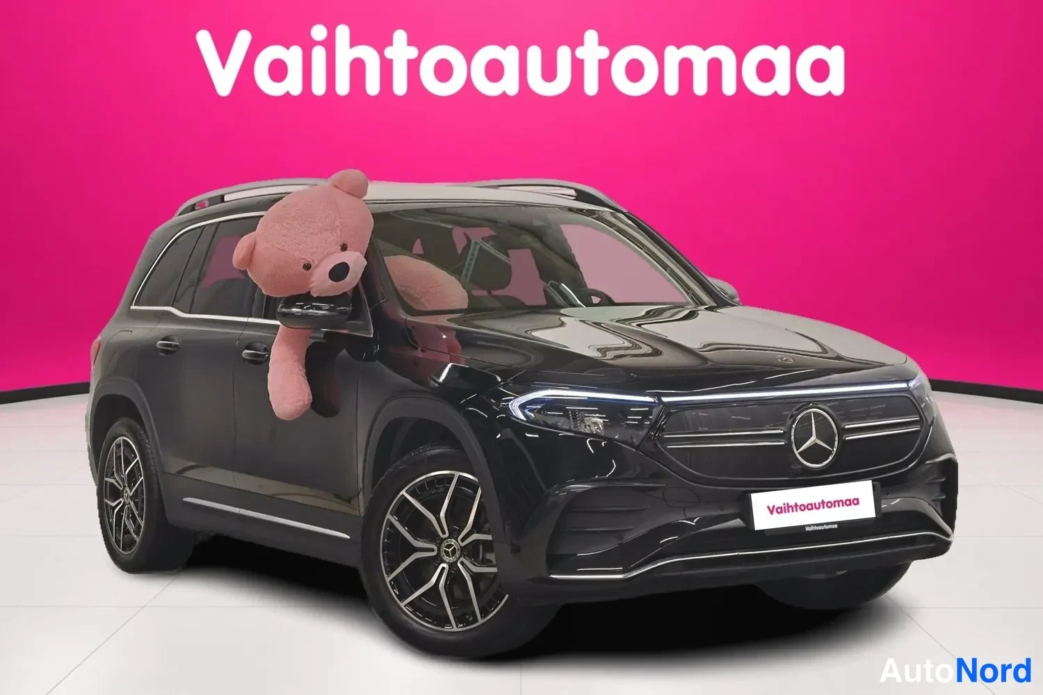 Etuviisto, nokka oikealle: Musta Mercedes-Benz EQB etu- ja sivulta oikeasta kulmasta, keulalla Mercedes-tähti ja LED-päiväajovalot, pinkki tausta.