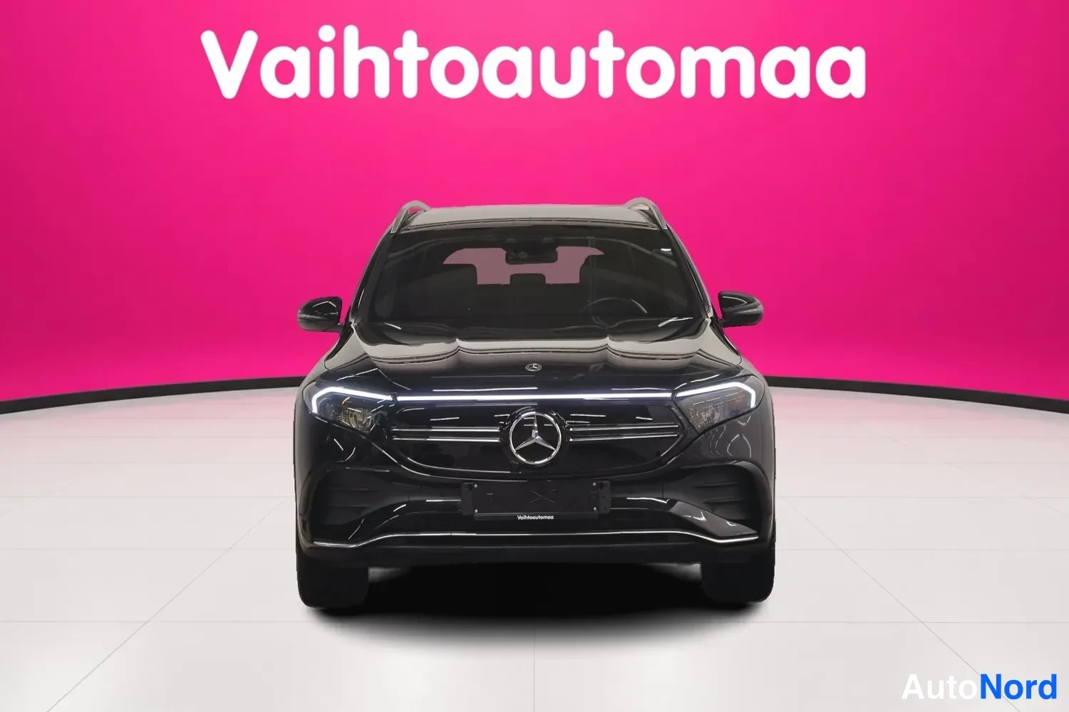 Edestä: Musta Mercedes-Benz EQB suoraan edestä, Mercedes-tähti keskellä etusäleikköä ja LED-ajovalot, pinkki tausta.
