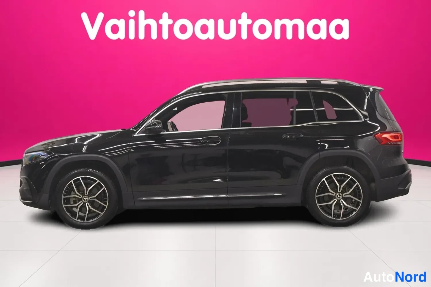 Etuviisto, nokka vasemmalle: Musta Mercedes-Benz EQB vasemmalta sivulta, kattokaiteet, moniportsaavantelevat kevytmetallivanteet ja tummat takaovien lasit, pinkki tausta.