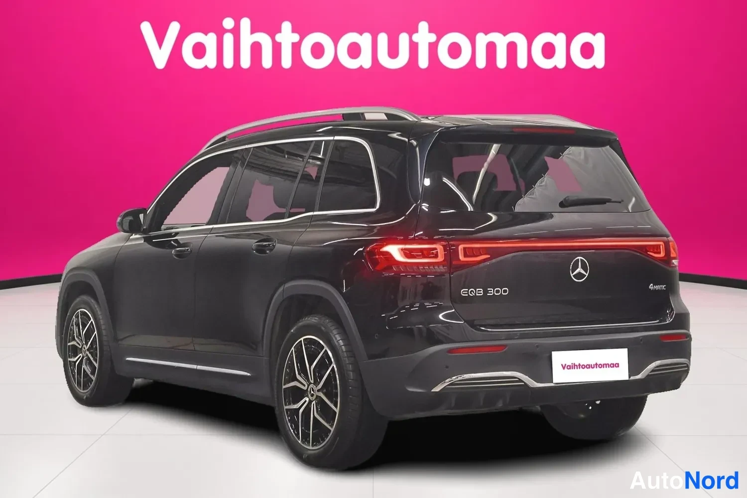 Takaviisto: Mustan Mercedes-Benz EQB:n takakulmanäkyvää: LI-tyylinen takavalojie, neljä pyörää ja kattotelineet näkyvät pinkkiseinämää vasten.