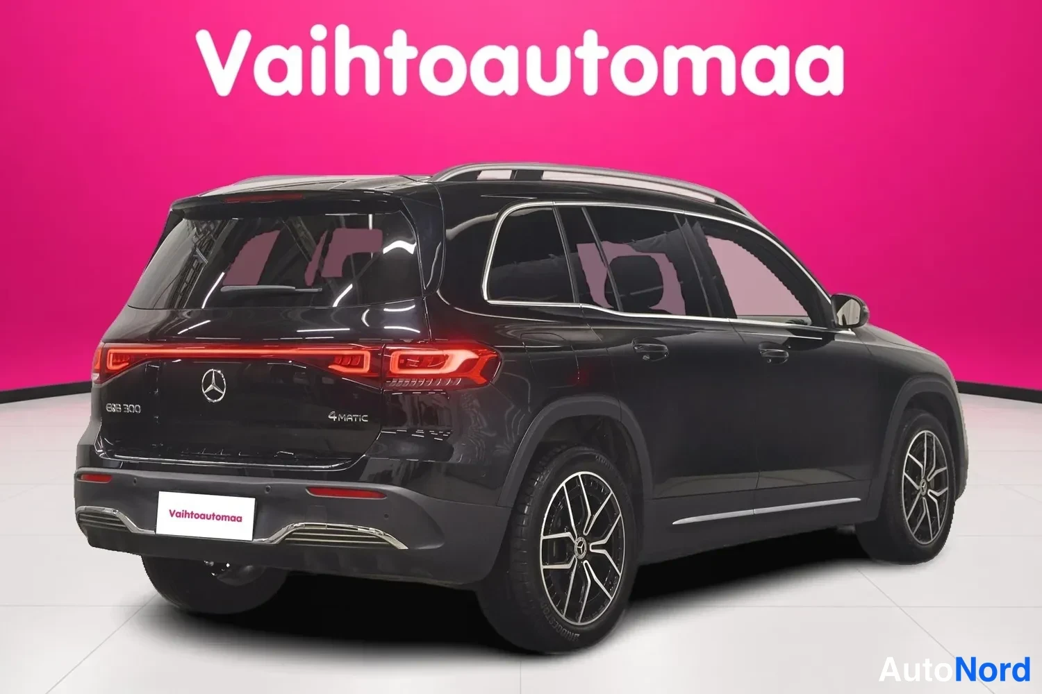 Takaviisto: Mustan Mercedes-Benz EQB:n takakulmanäkymä vasemmalta: kattotelineet, LI-muotoinen takavalo ja alumiinivanne näkyvät pinkkiseinämää vasten.
