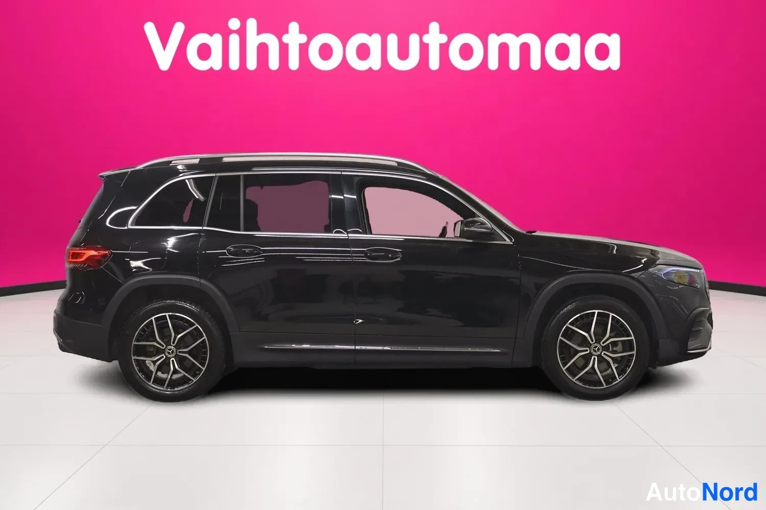 Sivulta: Mustan Mercedes-Benz EQB:n sivunäkymä: pitkä akseliväli, kattotelineet ja kaksivärinen alumiinivannet näkyvät pinkkiseinämää vasten.