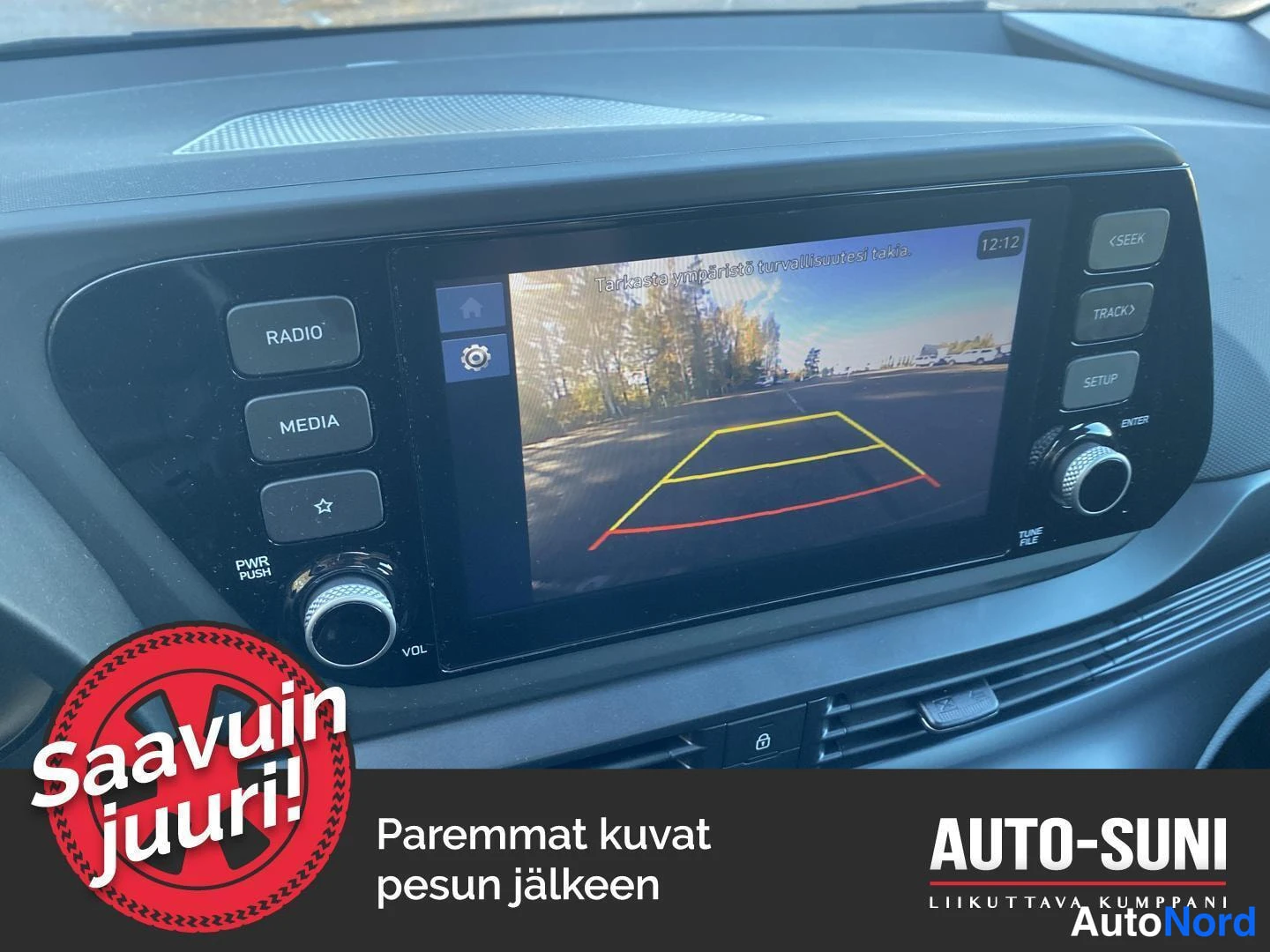 Edestä: Hyundai i20 - Kojelaudan infotainment-näyttö, jolla näkyy peruutuskameran kuva ohjausviivoineen ja teksti 'Tarkasta ympäristö turvallisuutesi takia'.