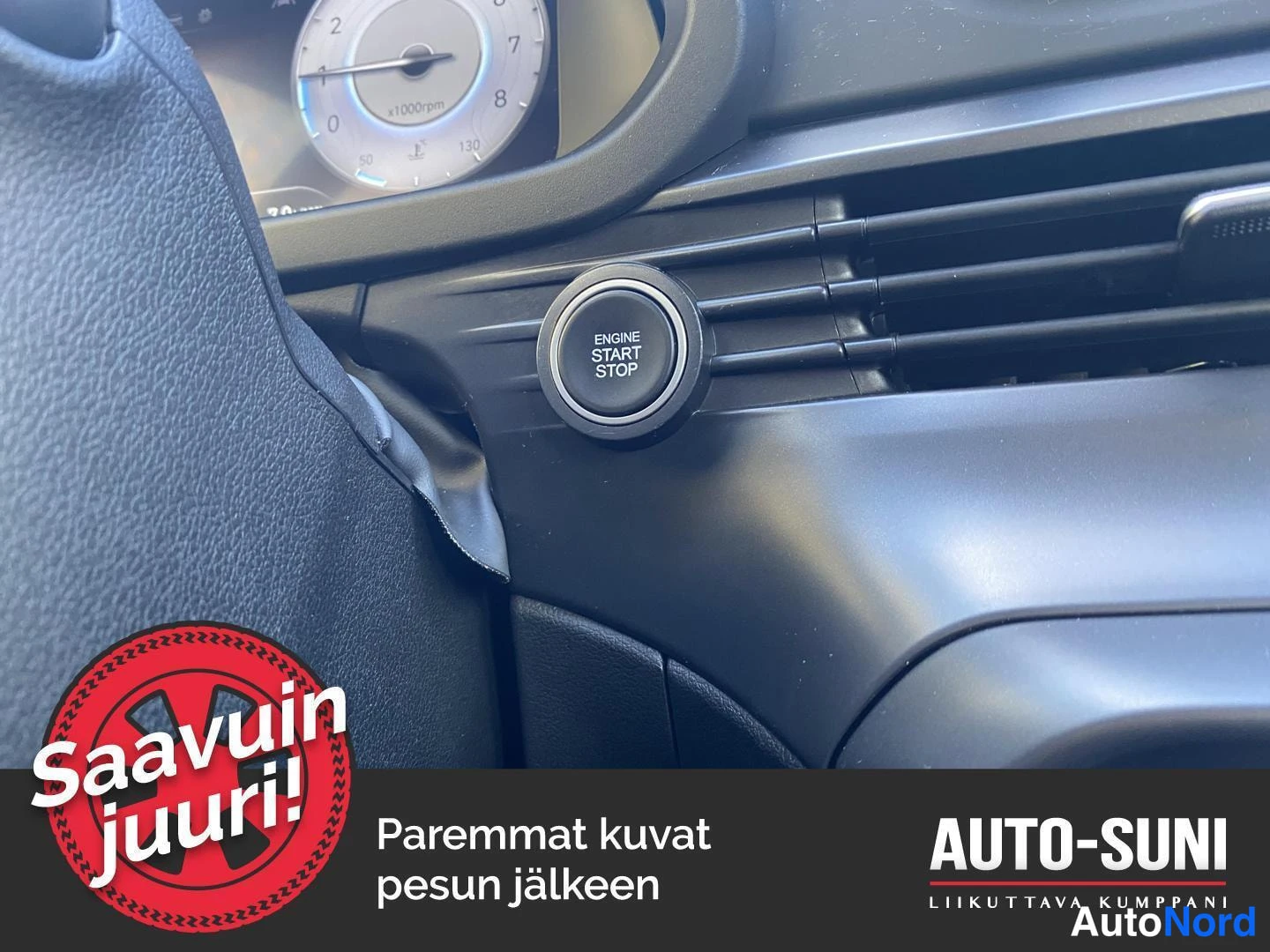 Yksityiskohta: Kojelaudan ajokytkin nappi, johon on painettu ENGINE START STOP, ja osa mittaristosta.