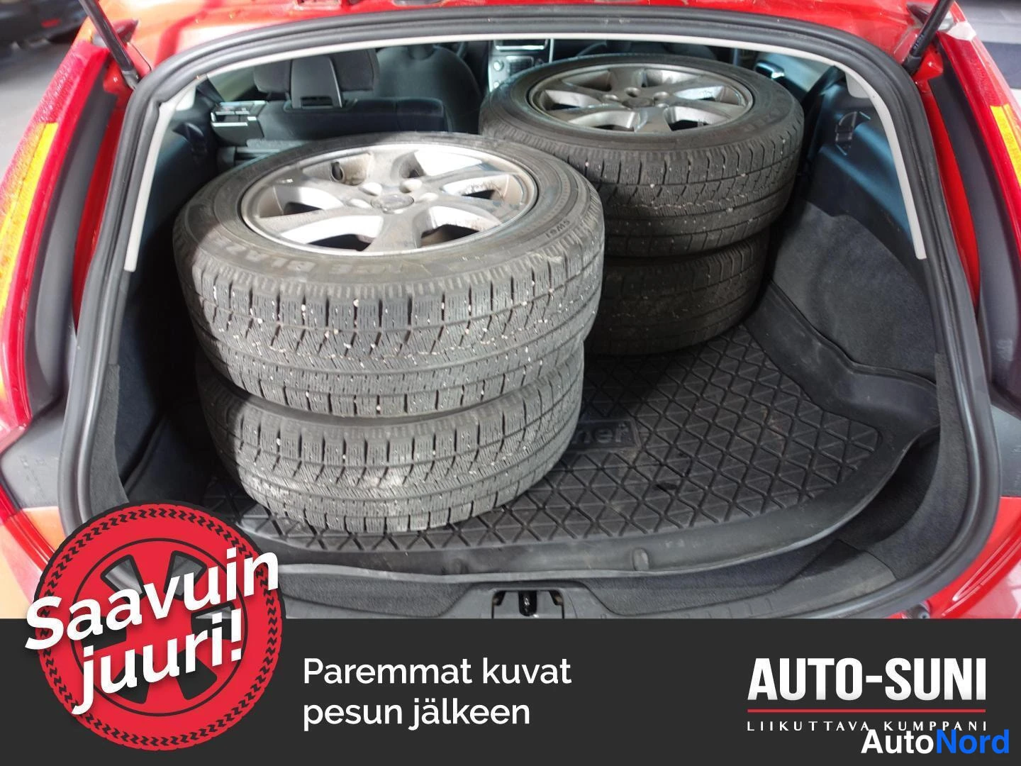 Edestä: Auton takaluukku auki: neljarenkainen talvirenkaset vanteineen lavetilla, taustapenkin nojaimet nakyvat.