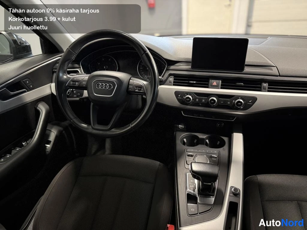 Muu: Auto interior: ohjauspyörä Audi-merkillä, kojelauta, keskikonsoli vaihteenvalitsimella, yläosassa tekstiä tarjouksesta.