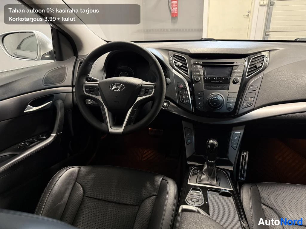 Muu: Hyundai I40:n etuosa sisältä: Hyundai-ohjauspyörä, tumma kojelauta, radiopaneeli ja vaihteenvalitsin.
