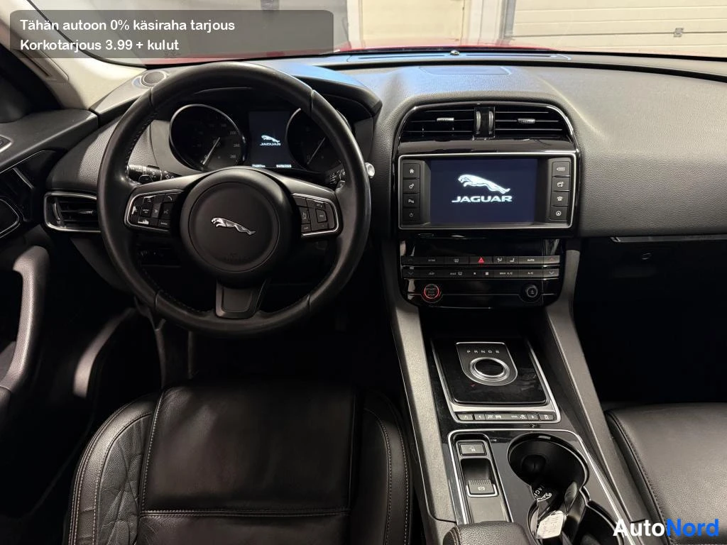 Edestä: Auton kojelauta ja ohjaustehostin Jaguar-logolla, infotainment-näyttö käynnissä.