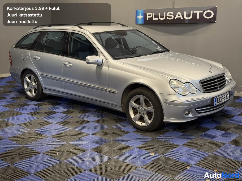 Etuviisto, nokka oikealle: Hopeinen Mercedes-Benz C 180 Kompressor -farmari etuokolänäkymästä sisätiloissa, rekisterikilpi, kattokaiteet ja alumiinivanteet näkyvissä.