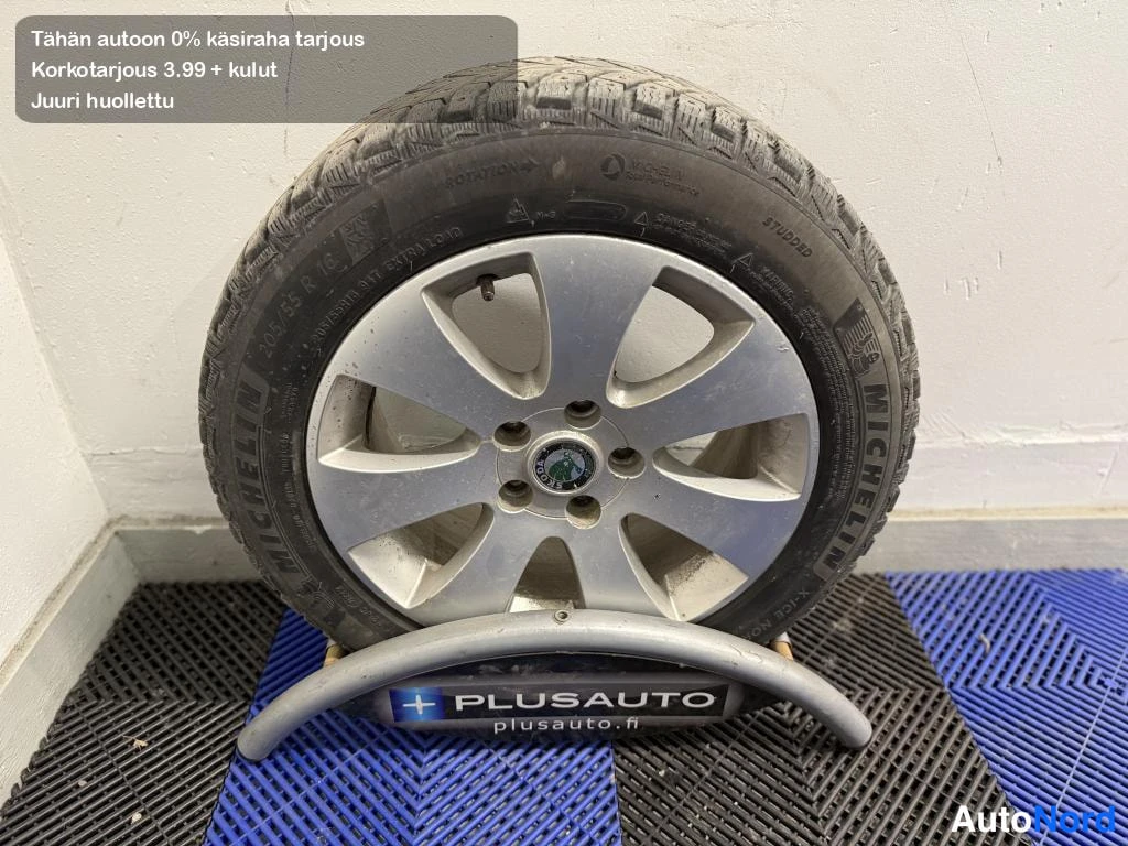 Yksityiskohta: Kevätrengas Michelin-renkaalla hopeavanteella seisoo telineessä PLUSAUTO-logolla lattiamatolla.