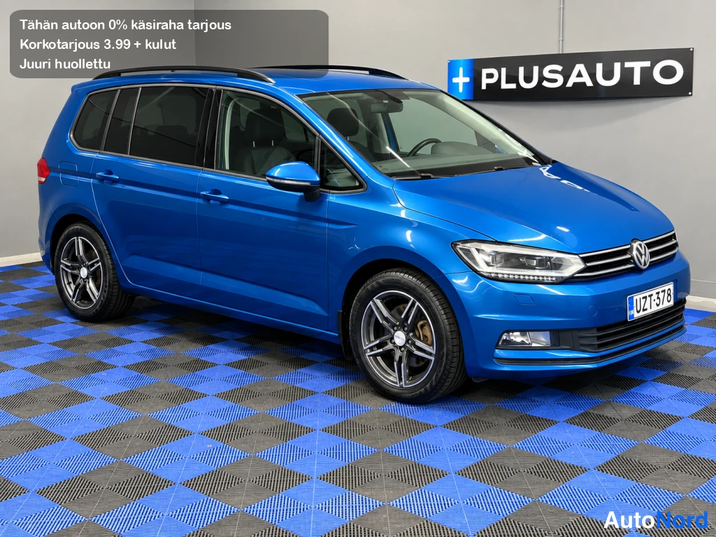 Etuviisto, nokka oikealle: Sininen Volkswagen Touran -henkilöauto etukulmasta oikealta kuvattuna sisätiloissa.