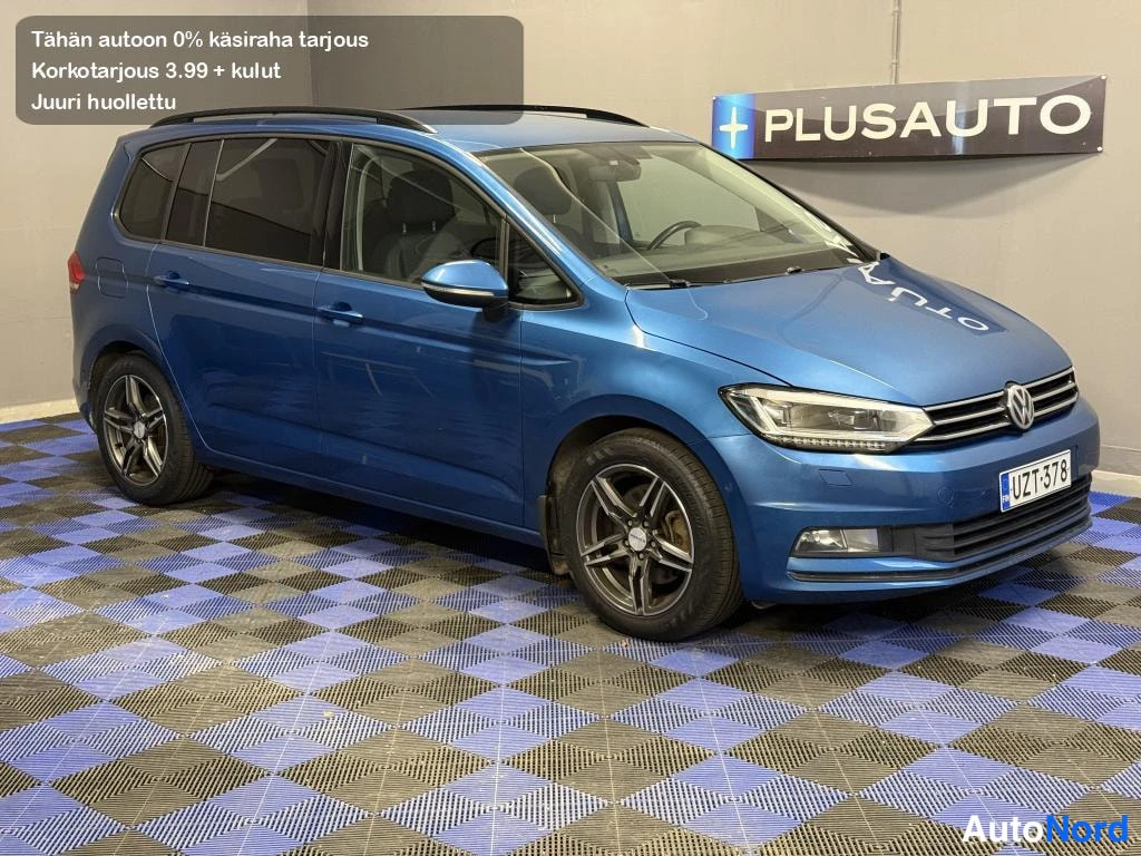 Etuviisto, nokka oikealle: Sininen Volkswagen Touran -henkilöauto etukulmasta oikealta kuvattuna sisätiloissa.