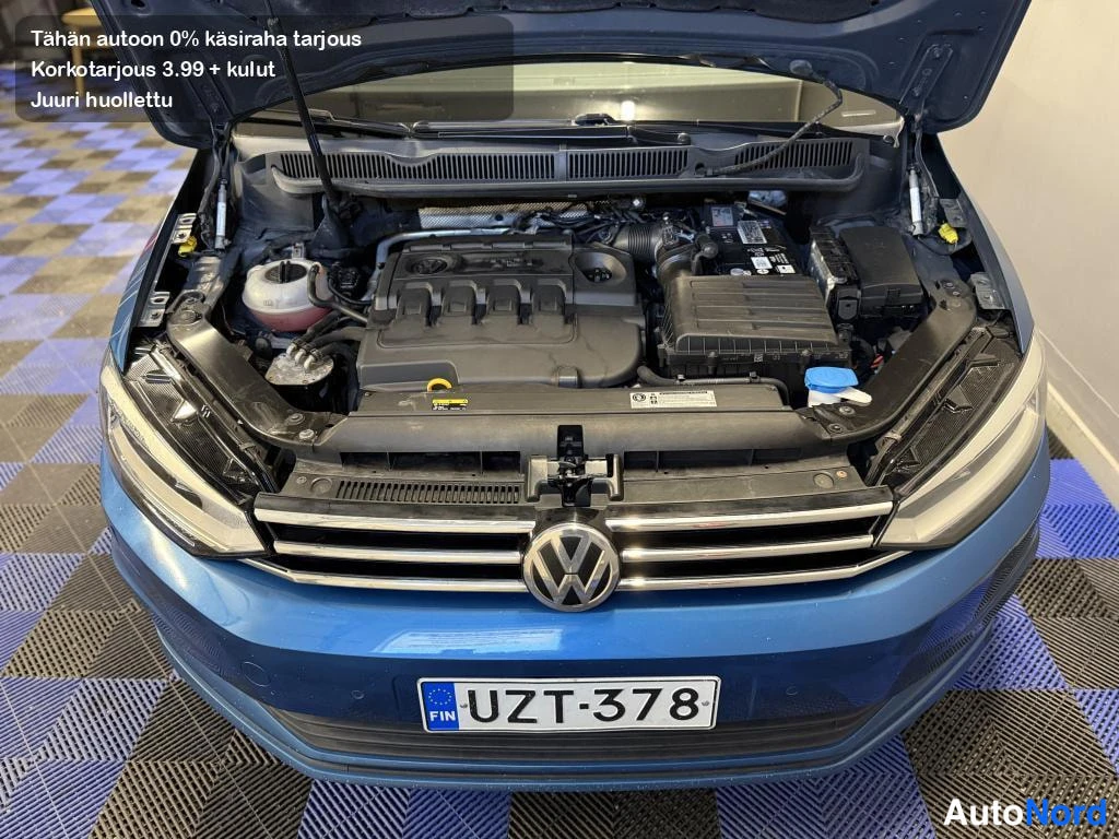 Edestä: Sinisen Volkswagenin konehuone auki edestä, moottori, jäähdytinaineen täyttöaukko ja akku näkyvät, etupuskurissa rekisterikilpi.