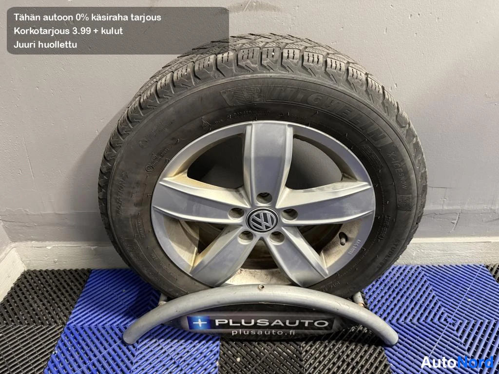 Yksityiskohta: Vaihtopyörä telineessä: hopeisen viisikanta-vanteen VW-merkki ja Michelin-kitkarengas, telineessä PLUSAUTO-logo.