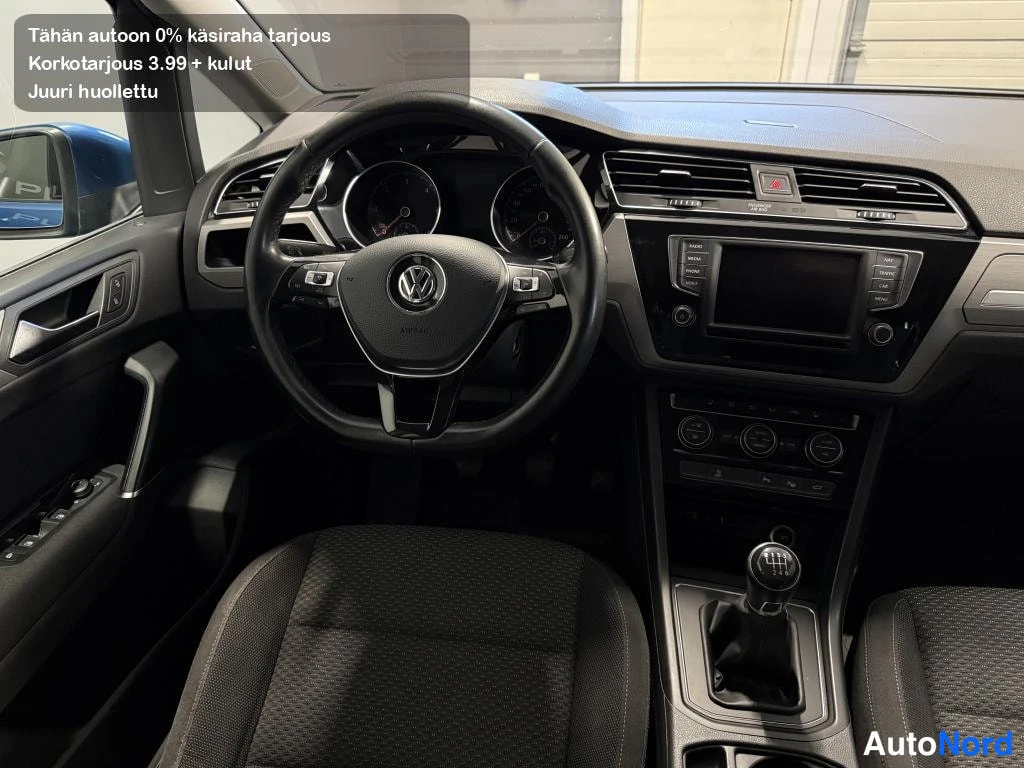 Muu: Volkswagenin ohjaamo: VW-merkitty ohjauspyörä, manuaalivaihtaja, infotaustanappi ja keskikonsoli.