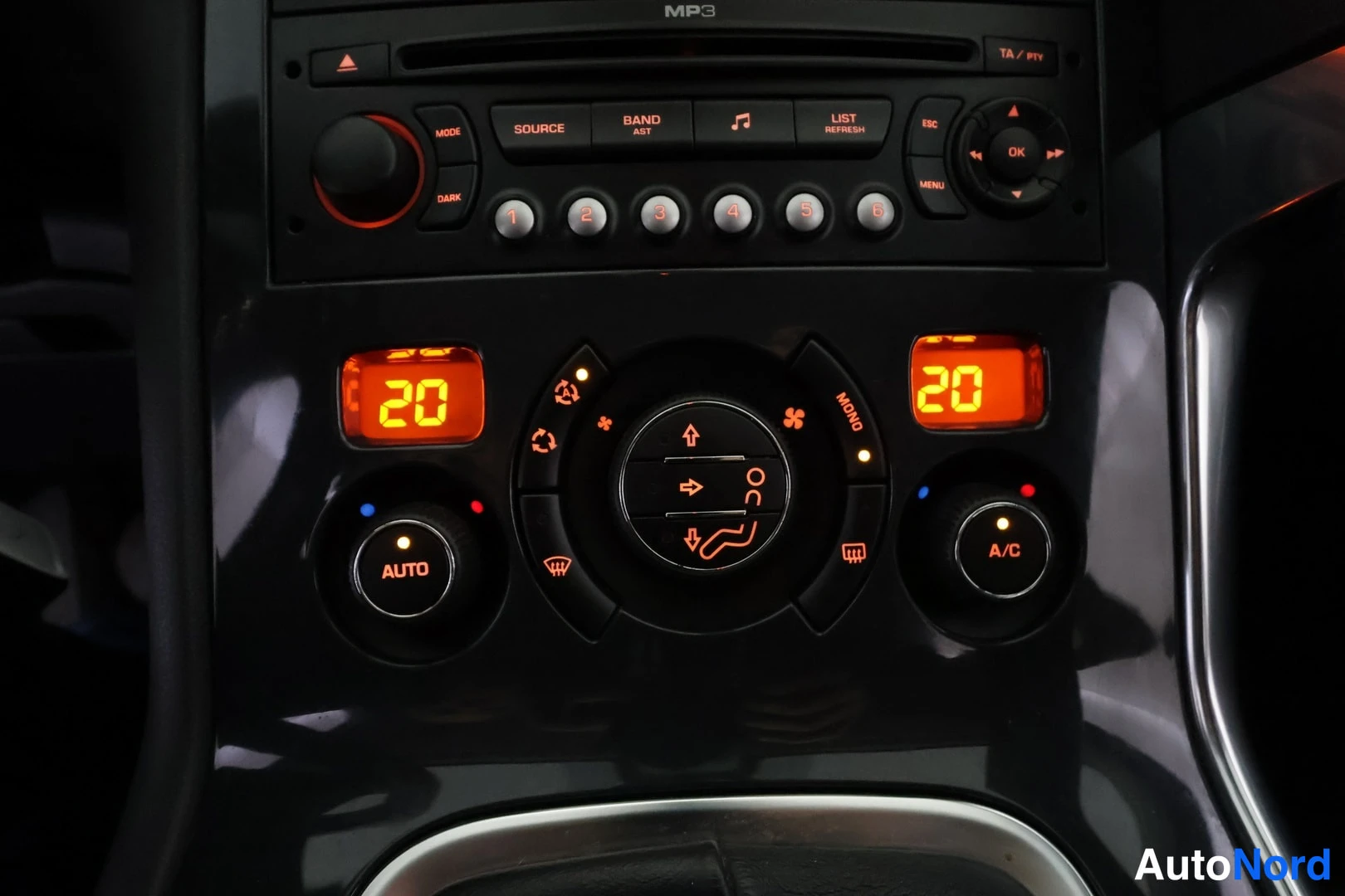 Muu: Lähikuva keskikonsolin ilmastoinnista: AUTO- ja A/C-säätimet, kaksialueiset 20 asteen näytöt ja radiopaneeli ylhäällä.