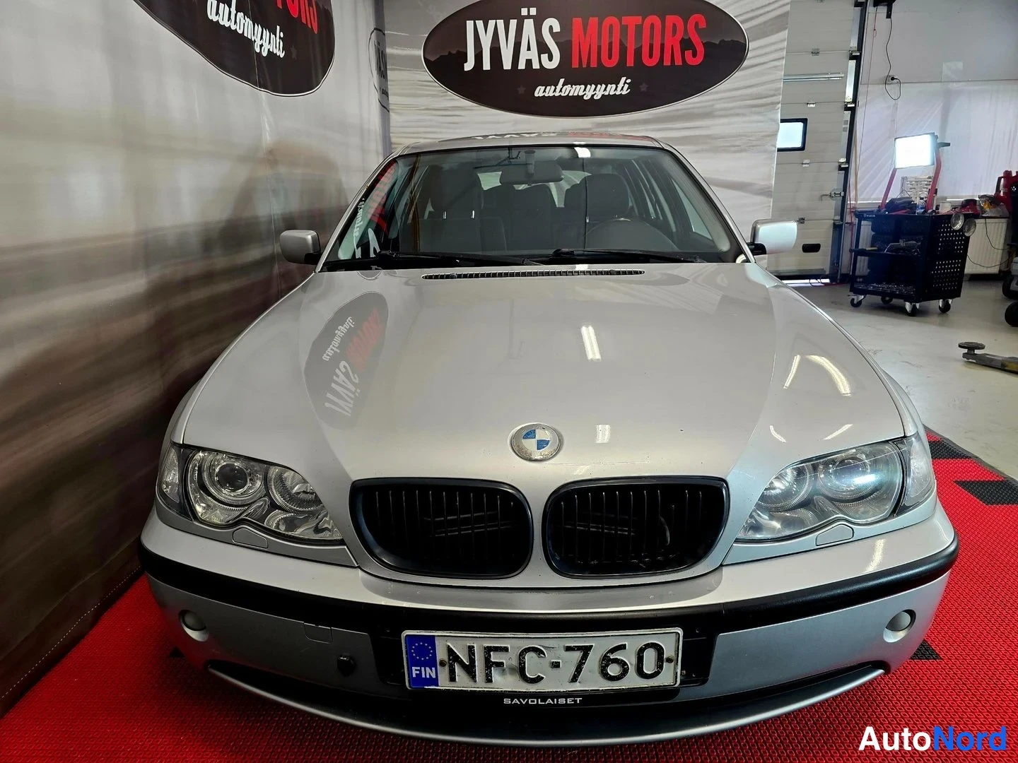 Edestä: Hopeanharmaan auton etupää showroomissa, rekisterikilpi, Jyväs Motors -taustabanneri.