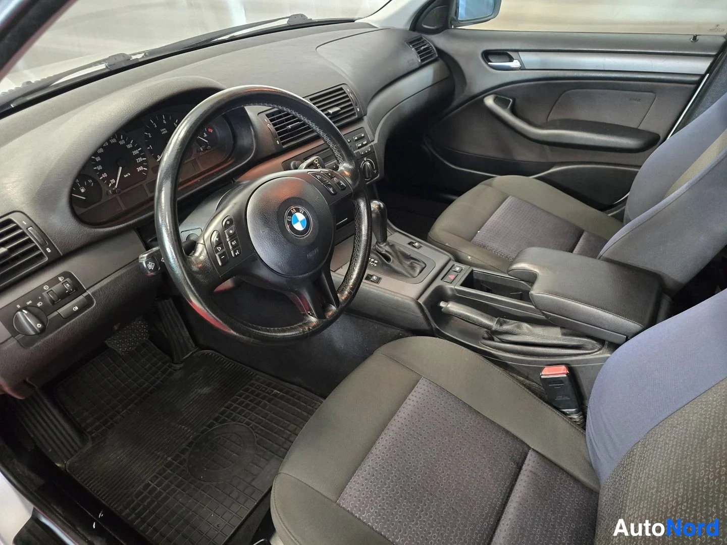Muu: Matkustamon näkymä kuljettajan ovesta: kaksi etuistuinta, BMW-ohjauspyörä, käsijarru ja tummat kangasistuimet.