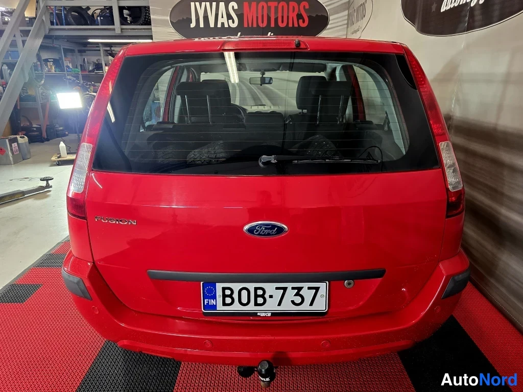 Takaa: Punaisen Ford Fusion -tilan takaosa: takanapit, Ford-merkki ja FUSION-teksti, rekisterikilpi ja takavaijerin kiinnitys; kuva sisätiloissa.